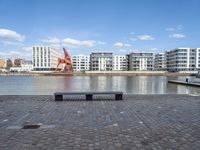 an empty bench sitting on the side of a river with buildings in the background and water behind