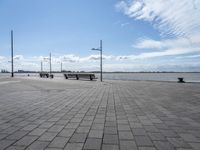 several benches are standing on the stone area near the ocean under a sky with white clouds