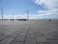several benches are standing on the stone area near the ocean under a sky with white clouds