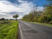 a long asphalt road leads to a green field with trees and bushes, as clouds float overhead