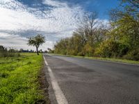 a long asphalt road leads to a green field with trees and bushes, as clouds float overhead