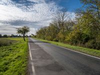 a long asphalt road leads to a green field with trees and bushes, as clouds float overhead