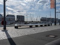 the bench is on a brick area near the water and buildings with sailboats docked in the background