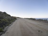 a lone dirt road is shown with blue skies in the background of the picture a hill with bushes