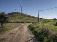 a dirt road going up to a grassy hill near windmills and power lines on a hillside