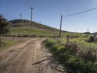 a dirt road going up to a grassy hill near windmills and power lines on a hillside