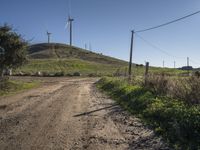 a dirt road going up to a grassy hill near windmills and power lines on a hillside