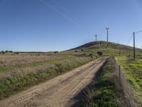 a dirt road passes by several wind turbines on the hillside above the road to the right of it