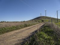 a dirt road passes by several wind turbines on the hillside above the road to the right of it