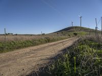 a dirt road passes by several wind turbines on the hillside above the road to the right of it