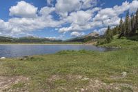 there is a lake surrounded by some trees and grass near some trees and mountains in the background