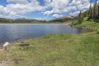 there is a lake surrounded by some trees and grass near some trees and mountains in the background