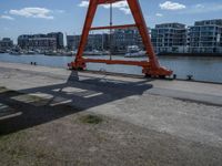 a large red crane in front of buildings on the river bank by the pier and boats