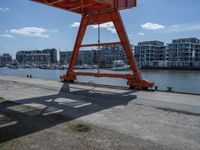 a large red crane in front of buildings on the river bank by the pier and boats