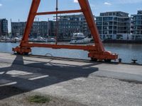 a large red crane in front of buildings on the river bank by the pier and boats