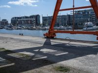 a large red crane in front of buildings on the river bank by the pier and boats