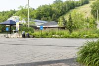 a person is walking on a sidewalk, while an umbrella stands in the background and shrubs