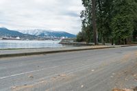 an empty street is next to a waterfront with mountains in the background with yellow leaves