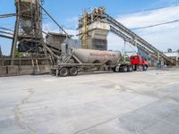 a red dump truck is parked near concrete mills and grain silos in the background