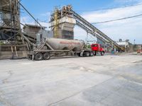 a red dump truck is parked near concrete mills and grain silos in the background