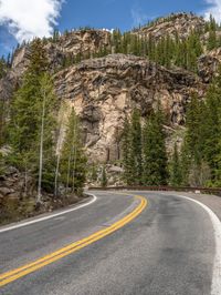 the curved road curves in front of a mountain area with pine trees behind it,