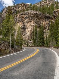 the curved road curves in front of a mountain area with pine trees behind it,