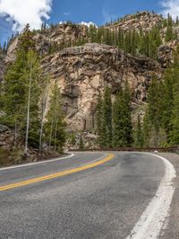 the curved road curves in front of a mountain area with pine trees behind it,