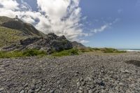 the hill has rocks in front of the ocean with clouds overhead and grass below it