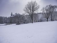 a man walking across snow covered ground in park area area with buildings in background and trees without leaves