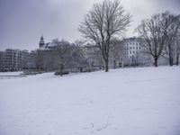 a man walking across snow covered ground in park area area with buildings in background and trees without leaves