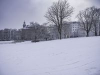 a man walking across snow covered ground in park area area with buildings in background and trees without leaves