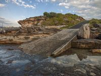 a concrete walkway across some rocks near water on a hill side with a large rock out in the distance