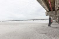 the underneath of an overpass overlooking the ocean at the beach's edge and the sand below