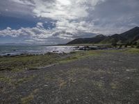 an empty beach with rocks and green grass on it on a cloudy day, with mountains in the background