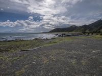 an empty beach with rocks and green grass on it on a cloudy day, with mountains in the background