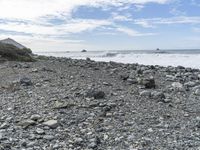 a black surfboard laying on a rocky beach and a wave coming in to shore