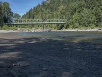 the view from the beach looking at a bridge with people on it above a shallow creek with a forested mountain in the background