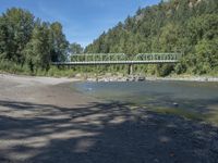 the view from the beach looking at a bridge with people on it above a shallow creek with a forested mountain in the background