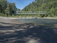 the view from the beach looking at a bridge with people on it above a shallow creek with a forested mountain in the background