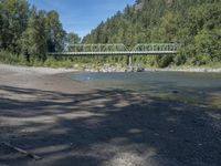 the view from the beach looking at a bridge with people on it above a shallow creek with a forested mountain in the background