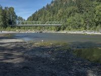 the view from the beach looking at a bridge with people on it above a shallow creek with a forested mountain in the background