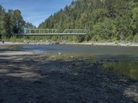 the view from the beach looking at a bridge with people on it above a shallow creek with a forested mountain in the background