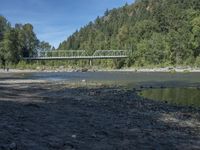 the view from the beach looking at a bridge with people on it above a shallow creek with a forested mountain in the background