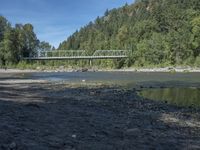 the view from the beach looking at a bridge with people on it above a shallow creek with a forested mountain in the background