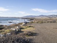 a beach scene, a bench and the ocean and mountains in the distance and blue skies