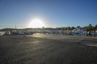 the sunset shines on some boats docked at the marina area, all near a fenced off parking space