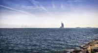 sail boats are seen sailing on the water near rocks and buildings in the background during sunny day