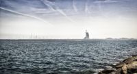 sail boats are seen sailing on the water near rocks and buildings in the background during sunny day