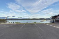 a parking lot is shown with water and a boat dock in the distance a red building and picnic tables sit on it