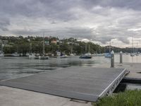 a pier with several boats parked along side the water and hills behind it below the clouds